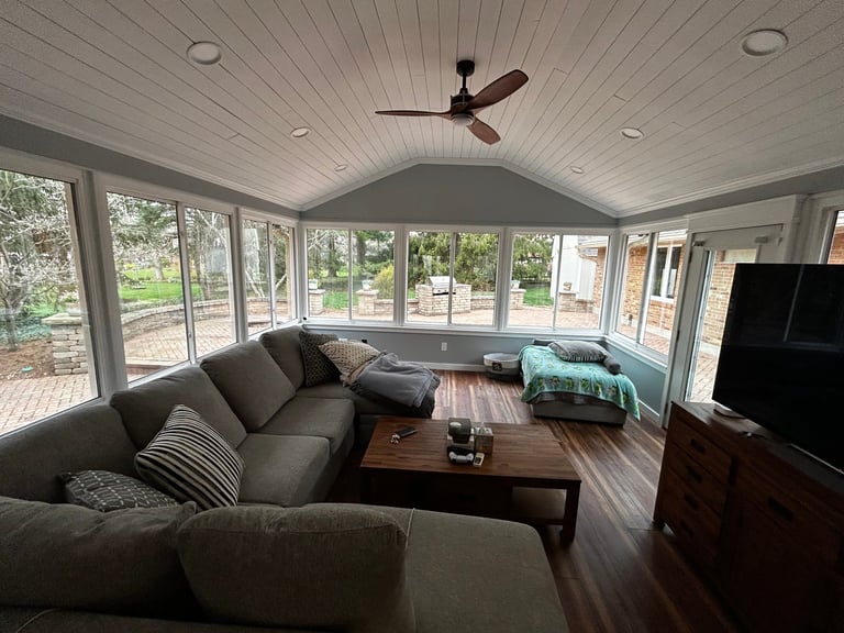Modern sunroom with gray couch, wooden coffee table, ceiling fan, and large windows overlooking garden