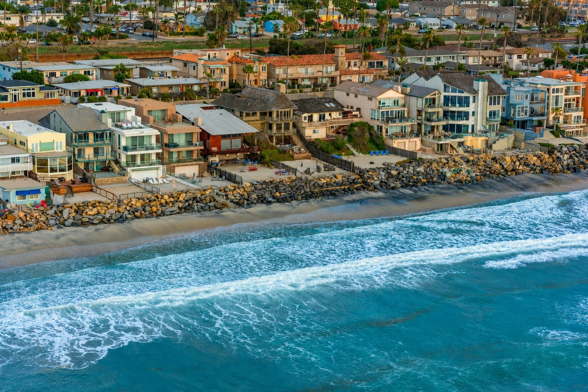 Aerial view of a colorful beachfront neighborhood with residential homes, sandy shore, and rolling ocean waves along the waterfront